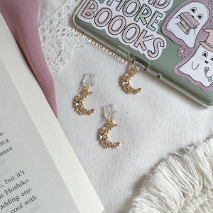 May include: Three gold crescent moon-shaped bookmarks with rhinestones. Each bookmark has a clear plastic clip to hold a page in a book.