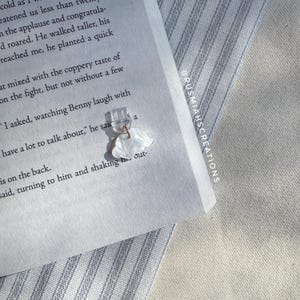 May include: A white, cloud-shaped earring with a clear plastic backing. The earring is on a book with text that reads "I asked, watching Benny laugh with a lot to talk about," he said, turning to him and shaking his out."