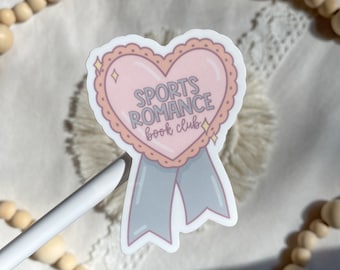 May include: A pink heart-shaped sticker with the text "SPORTS ROMANCE book club" inside. The heart has a white outline and a blue ribbon.