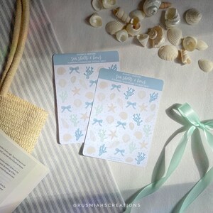 May include: Two sheets of stickers featuring illustrations of seashells, starfish, coral, and bows on a white background with blue accents. The sticker sheets are titled "Sea Shells & Bows" and are part of a planning/journaling sticker set.