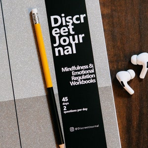 May include: A black and white journal with the text "Discreet Journal" and "Mindfulness & Emotional Regulation Workbooks" on the cover. The journal has a silver glitter cover and a yellow pencil is lying on top of it. The text on the cover also says "45 days, 2 questions per day" and "@DiscreetJournal".