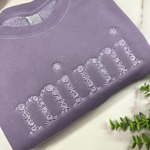 May include: A lavender sweatshirt with the embroidered word "mimi" in floral letters. The sweatshirt is made of a soft material and has a crew neck. The floral embroidery adds a delicate touch to the casual style.