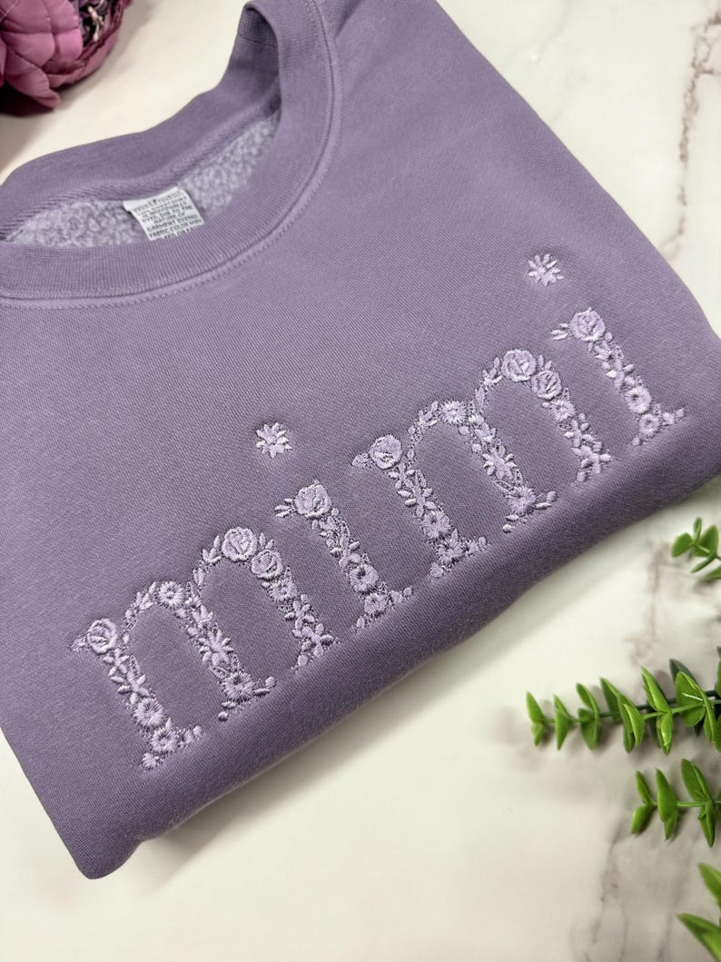 May include: A lavender sweatshirt with the embroidered word "mimi" in floral letters. The sweatshirt is made of a soft material and has a crew neck. The floral embroidery adds a delicate touch to the casual style.