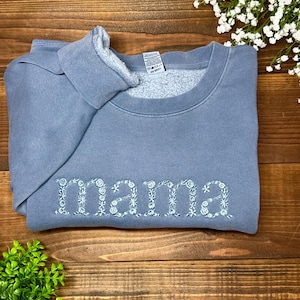 May include: A blue-grey sweatshirt with the word "mama" embroidered in white floral letters. The sweatshirt is folded on a wooden surface, with a small green plant and white flowers in the background.