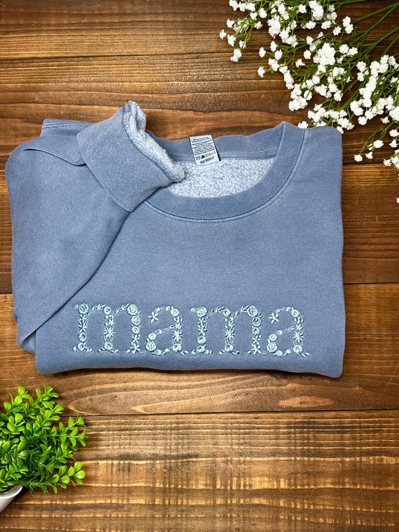 May include: A blue-grey sweatshirt with the word "mama" embroidered in white floral letters. The sweatshirt is folded on a wooden surface, with a small green plant and white flowers in the background.