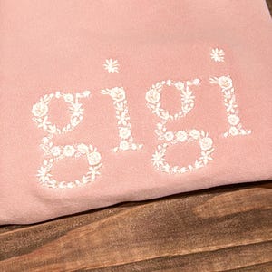 May include: A light pink sweatshirt with the name "Gigi" embroidered in white floral letters. The sweatshirt is made of a soft material and is displayed on a wooden surface. The floral embroidery adds a delicate touch to the casual style.