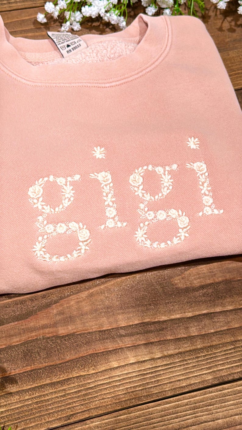 May include: A light pink sweatshirt with the name "Gigi" embroidered in white floral letters. The sweatshirt is made of a soft material and is displayed on a wooden surface. The floral embroidery adds a delicate touch to the casual style.