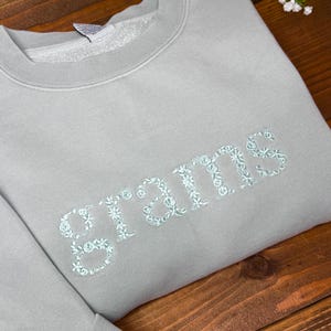 May include: A light gray sweatshirt with the word "grams" embroidered in a floral design. The sweatshirt is folded on a wooden surface, with white flowers in the background. The embroidery is a light blue color.