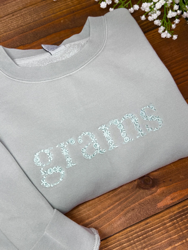 May include: A light gray sweatshirt with the word "grams" embroidered in a floral design. The sweatshirt is folded on a wooden surface, with white flowers in the background. The embroidery is a light blue color.