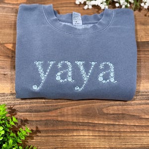 May include: A blue-grey sweatshirt with the word "YAYA" embroidered in light blue floral letters. The sweatshirt is folded and rests on a wooden surface, with greenery and white flowers in the background.