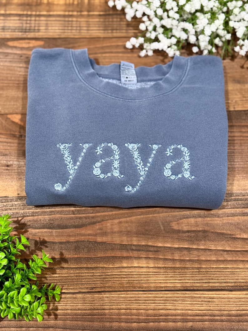 May include: A blue-grey sweatshirt with the word "YAYA" embroidered in light blue floral letters. The sweatshirt is folded and rests on a wooden surface, with greenery and white flowers in the background.