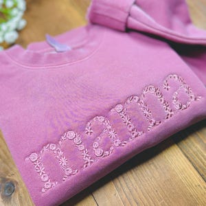 May include: A mauve-colored sweatshirt with the word "mama" embroidered in a floral design. The sweatshirt is folded on a wooden surface, with a matching folded sleeve visible. The floral embroidery adds a delicate touch to the garment.