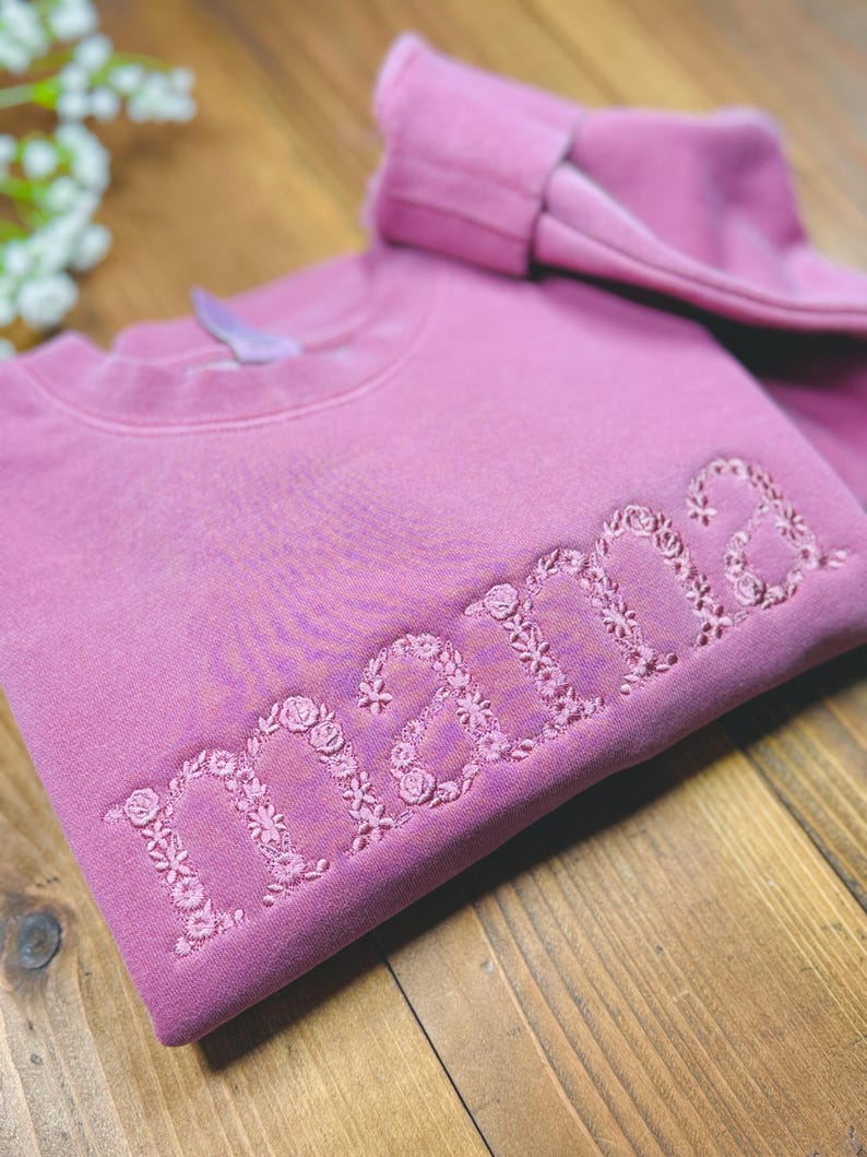 May include: A mauve-colored sweatshirt with the word "mama" embroidered in a floral design. The sweatshirt is folded on a wooden surface, with a matching folded sleeve visible. The floral embroidery adds a delicate touch to the garment.