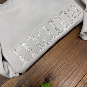 May include: A beige sweatshirt with the word "mama" embroidered in white floral letters. The sweatshirt is folded on a dark wooden surface, with a small green plant in the corner.