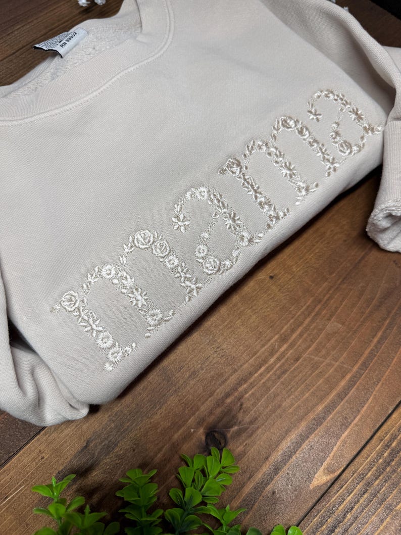 May include: A beige sweatshirt with the word "mama" embroidered in white floral letters. The sweatshirt is folded on a dark wooden surface, with a small green plant in the corner.