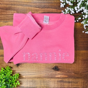 May include: A pink sweatshirt with the name "Mamie" embroidered in a floral design. The sweatshirt is folded on a wooden surface, with a small green plant and white flowers in the background.