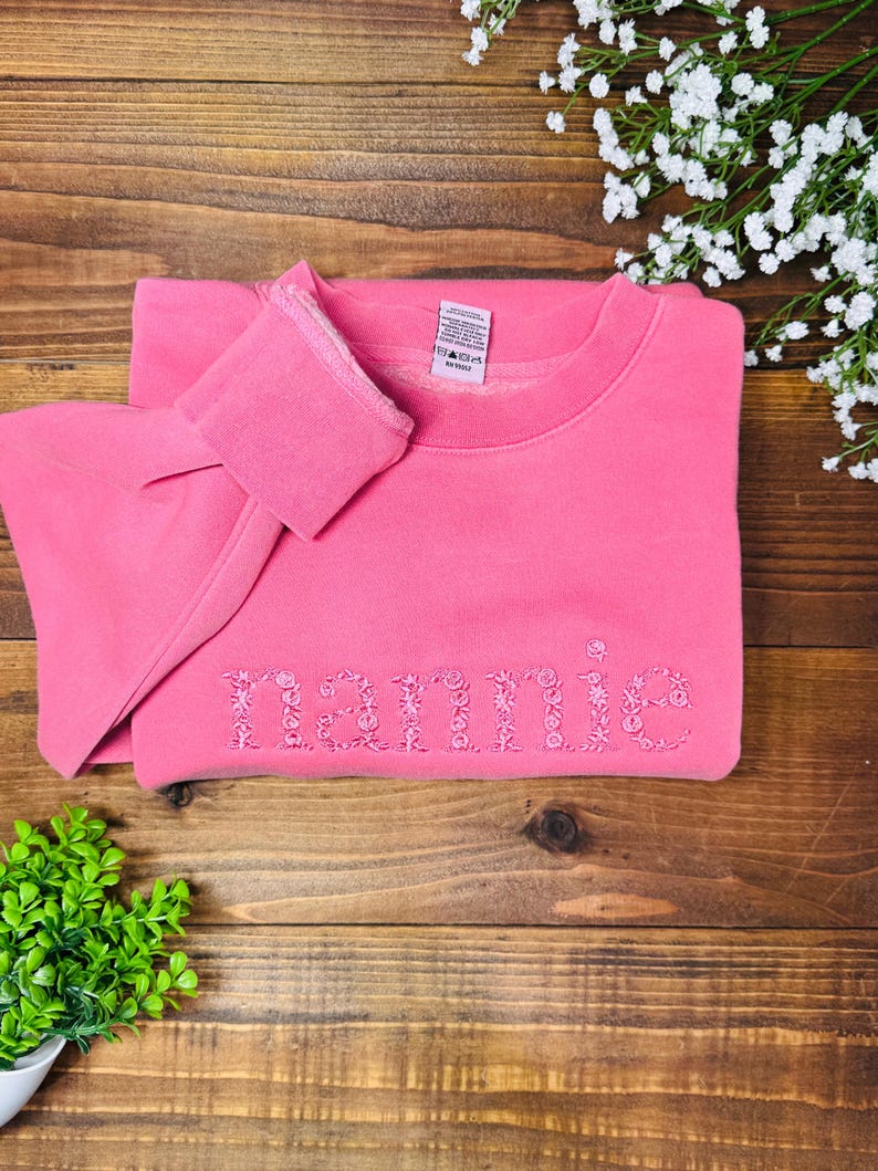 May include: A pink sweatshirt with the name "Mamie" embroidered in a floral design. The sweatshirt is folded on a wooden surface, with a small green plant and white flowers in the background.