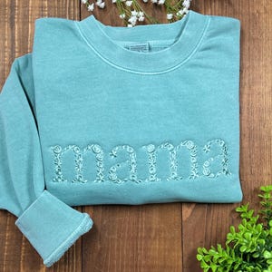 May include: A teal sweatshirt with the embroidered word "mama" in floral letters. The sweatshirt has a crew neck and ribbed cuffs. The garment is displayed on a wooden surface with floral accents.