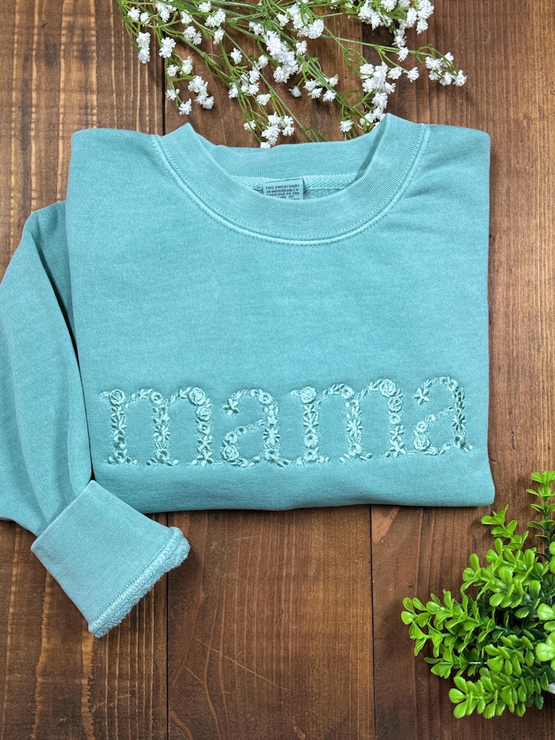 May include: A teal sweatshirt with the embroidered word "mama" in floral letters. The sweatshirt has a crew neck and ribbed cuffs. The garment is displayed on a wooden surface with floral accents.
