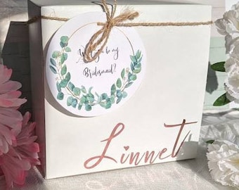 Może przedstawiać: Białe pudełko na prezent związane sznurkiem, z okrągłą etykietą z napisem "Will you be my Bridesmaid?" otoczoną zielonym wieńcem z eukaliptusa. Imię "Linnet" jest napisane kursywą na pudełku.
