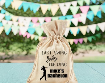 May include: A beige burlap drawstring bag with the text "LAST SWING Before THE RING mike's bachelor" in black. A silhouette of a golfer is also printed on the bag. The bag is sitting on a light blue surface with a blurred background of colorful triangular flags.