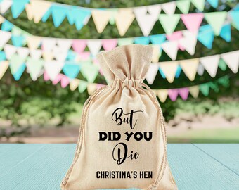 May include: A beige burlap drawstring bag with the text "But DID YOU Die CHRISTINA'S HEN" in black. The bag is tied at the top with a natural rope. The background features a blurred outdoor scene with colorful triangular flags.