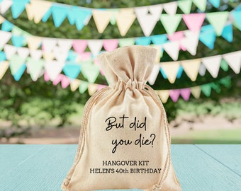May include: A beige fabric bag with the text "But did you die?" and "Hangover Kit Helen's 40th Birthday" printed on it. The bag is sitting on a light blue wooden surface with a blurry background of colorful triangle flags.