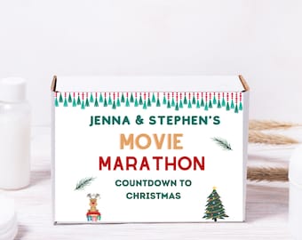 Op de afbeelding: Een witte doos met de tekst "Jenna & Stephen's Movie Marathon Countdown to Christmas". De doos heeft een feestelijk ontwerp met groene en rode kwastjes, een rendier en een kerstboomillustratie. De doos staat op een wit houten oppervlak.