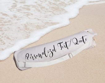 May include: A rolled-up, light-colored beach towel with a white strap, lying on a sandy beach with gentle waves. The towel has the words "Personalized Text / Quote" printed on it in black script.