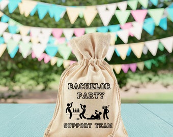 May include: A beige drawstring bag with the text "Bachelor Party Support Team" printed on it. The bag features a black graphic of three stick figures, one at a bar, one passed out on the floor, and one holding a bottle.