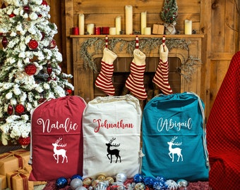 May include: Three personalized Christmas drawstring bags in red, white, and teal. Each bag features a name in script and a white deer silhouette. The bags are set against a festive holiday backdrop with a Christmas tree and stockings.