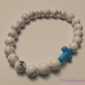 May include: White stone bead bracelet with a light blue cross charm.
