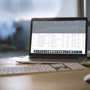 May include: A laptop computer with a white keyboard and a mouse on a wooden desk. The laptop screen displays a spreadsheet with a line graph showing an upward trend. The graph is labeled "Revenue" and the spreadsheet shows data for "Date", "Source", "Orders", "Revenue", "Change in Revenue", "% Change", and "Attribution".