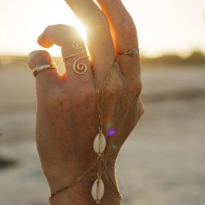 May include: A gold-toned hand chain featuring two white cowrie shells connected by delicate metal links. The jewelry piece extends from a wrist chain to a central ring attachment, complemented by three additional gold-toned rings on the fingers.