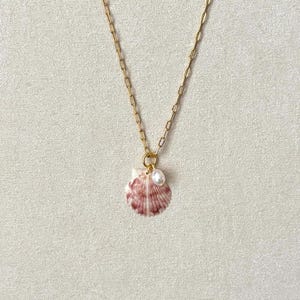 May include: A gold-colored chain necklace with a pendant featuring a small, pink and white seashell. A small white pearl is attached to the seashell. The necklace is displayed on a neutral background.
