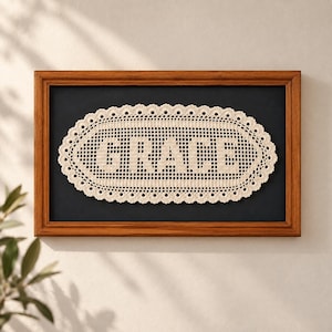 May include: An oval-shaped cream-colored crochet doily featuring the word GRACE in a grid-like pattern, framed in a rectangular wooden frame with a solid black background. The doily has a scalloped edge and a detailed open-work stitch design.