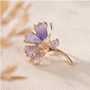 May include: A gold-toned brooch shaped like a ginkgo leaf, featuring translucent light purple enamel petals with gold edges and small clear stones embedded near the center stem, creating a delicate and detailed botanical accessory.
