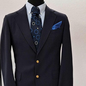 May include: Navy blue blazer with gold buttons, paired with a light blue button-down shirt and a patterned tie. A blue pocket square adds a pop of color. The blazer is displayed on a mannequin.