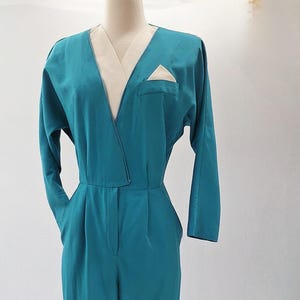 May include: Teal jumpsuit with a V-neck and long sleeves. It features a cream-coloured lapel and pocket detail. The waist is cinched, and the trousers have a wide-leg design. The garment is displayed on a mannequin.