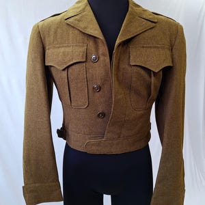 May include: A vintage olive-drab jacket with a tailored fit. The jacket features two chest pockets, button closures, and epaulettes. The sleeves have buttoned cuffs. The jacket is displayed on a mannequin against a white backdrop.