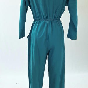 May include: Teal jumpsuit featuring long sleeves and a V-neck. The waist is gathered with an elasticated band, and the wide-leg trousers create a flowing silhouette. The back view highlights the garment's design and fit.