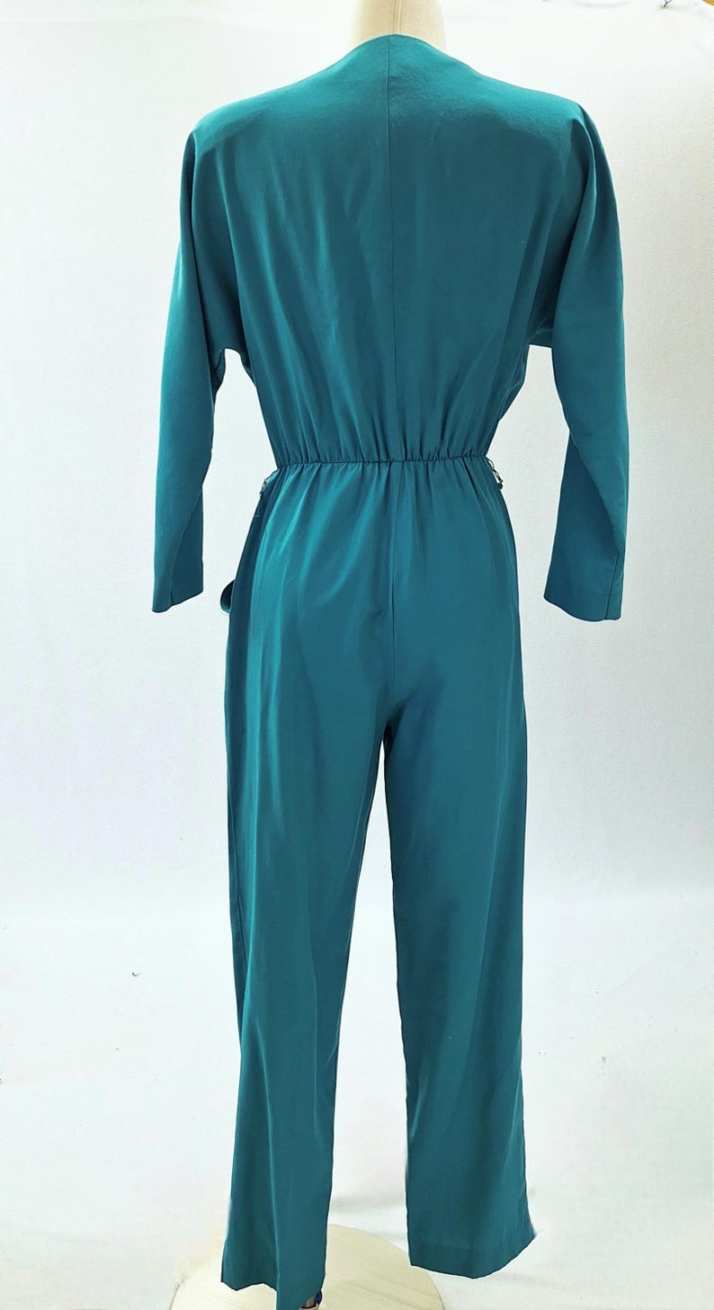 May include: Teal jumpsuit featuring long sleeves and a V-neck. The waist is gathered with an elasticated band, and the wide-leg trousers create a flowing silhouette. The back view highlights the garment's design and fit.