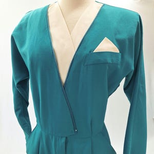May include: Teal jumpsuit with long sleeves and a V-neck. The neckline and pocket have a cream-coloured contrast. A cream pocket square is visible in the chest pocket. The jumpsuit has a tailored fit.