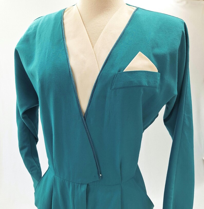 May include: Teal jumpsuit with long sleeves and a V-neck. The neckline and pocket have a cream-coloured contrast. A cream pocket square is visible in the chest pocket. The jumpsuit has a tailored fit.