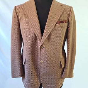 May include: A tan and brown herringbone patterned blazer featuring a notched lapel, two front buttons, and a chest pocket containing a dark red pocket square. The jacket has long sleeves and two flap pockets on the lower front section.