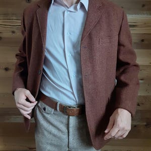 May include: A brown tweed blazer with a three-button closure. The blazer has a herringbone pattern and is worn over a light blue button-down shirt and beige pants.