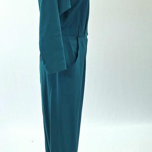 May include: Teal jumpsuit with long sleeves and a V-neck. The one-piece garment has a tailored fit with a defined waist and pockets. The jumpsuit is displayed on a mannequin against a white background.