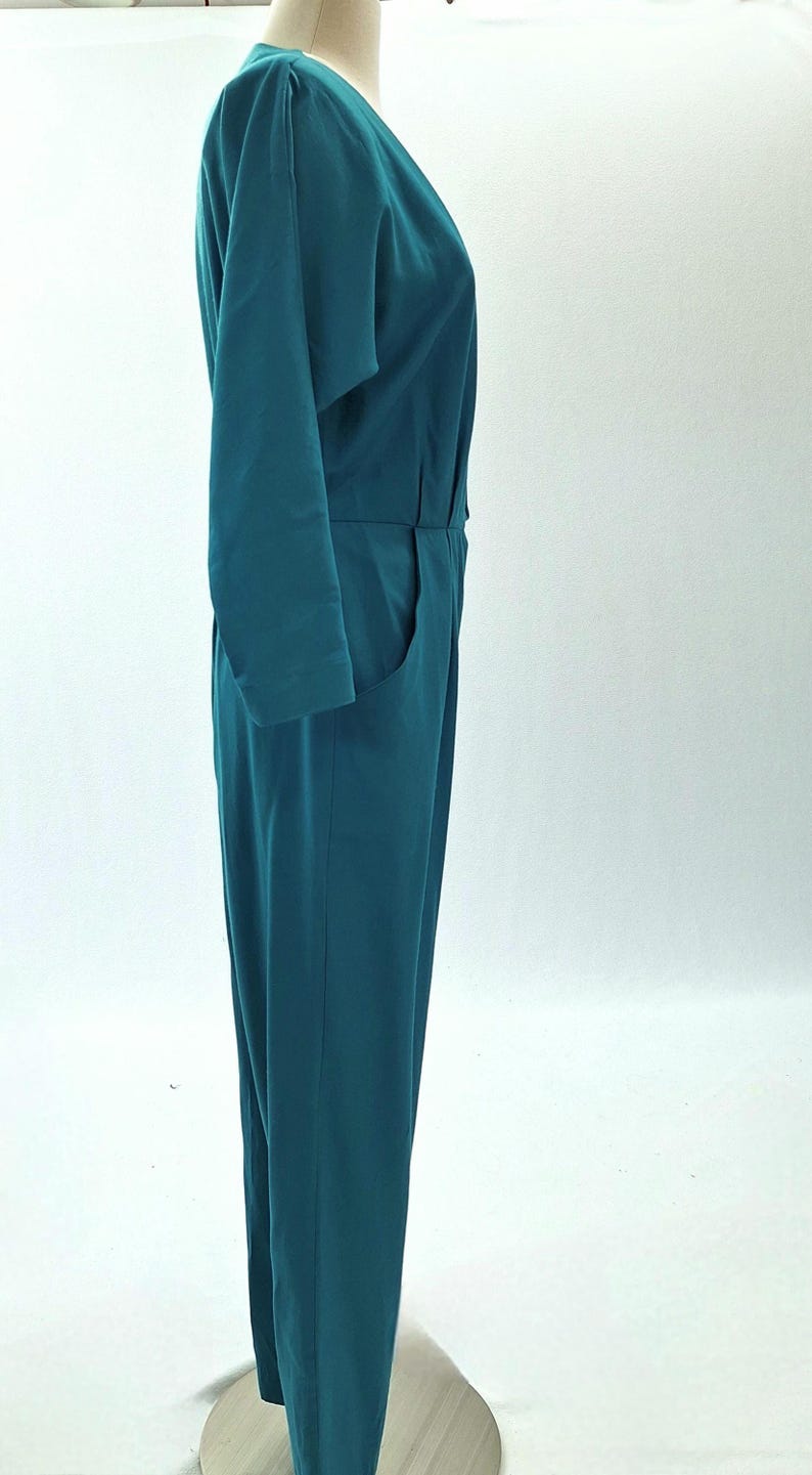 May include: Teal jumpsuit with long sleeves and a V-neck. The one-piece garment has a tailored fit with a defined waist and pockets. The jumpsuit is displayed on a mannequin against a white background.