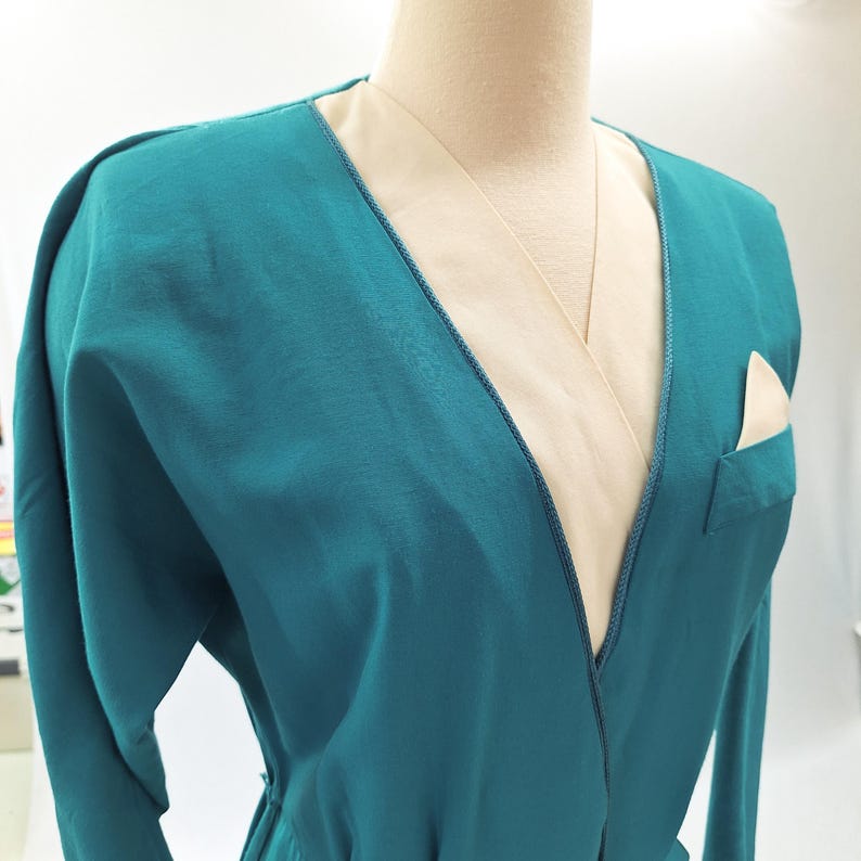 May include: Teal wrap dress with a beige lining and a pocket. The dress has long sleeves and a V-neck. The neckline and pocket are trimmed with darker teal stitching. The dress is displayed on a mannequin. The dress is a stylish garment.