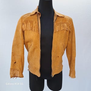 May include: A brown suede jacket with fringe detailing on the collar, cuffs, and front. The jacket has a zipper closure and two pockets.